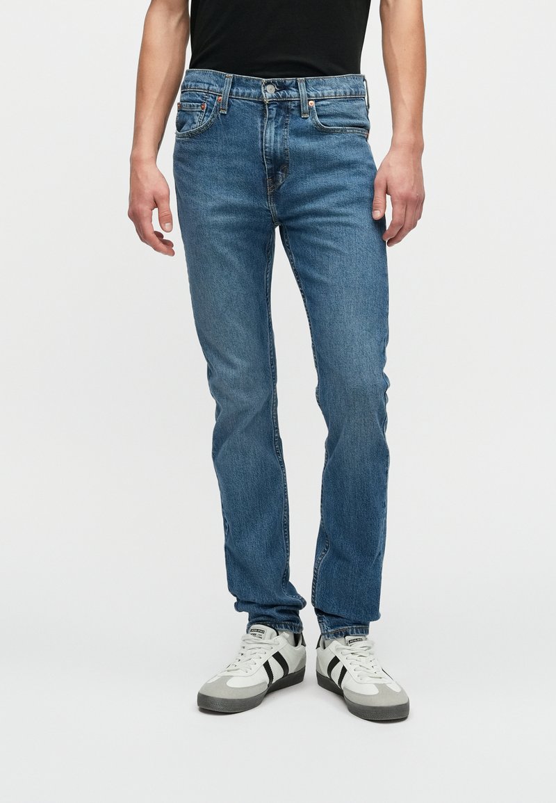 Pessoa vestindo jeans justos azuis e ténis brancos com riscas pretas, de pé contra um fundo claro e liso.