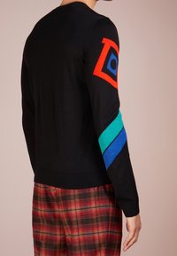 Zwarte trui met kleurrijke geometrische patronen in rood, blauw en groen op de mouwen. Voorzien van geribbelde boorden en zoom. Draagt met een geruite broek.