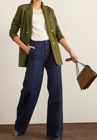 Olijfgroene dubbelbreasted blazer, witte T-shirt met borstzak, hoogwater donkere spijkerbroek met wijde pijpen en een bruine handtas met kettingen.