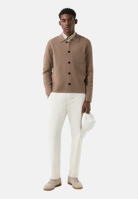 Braunes Hemd zum Knöpfen mit Tasche, darunter ein weißes Hemd mit Kragen, weiße Hose, beige Turnschuhe und eine weiße Kappe in der Hand gehalten.