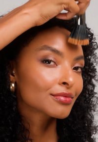 Femme aux cheveux bouclés appliquant du fond de teint sur le front avec un pinceau éventail, portant des boucles d'oreilles créoles dorées et un maquillage naturel.