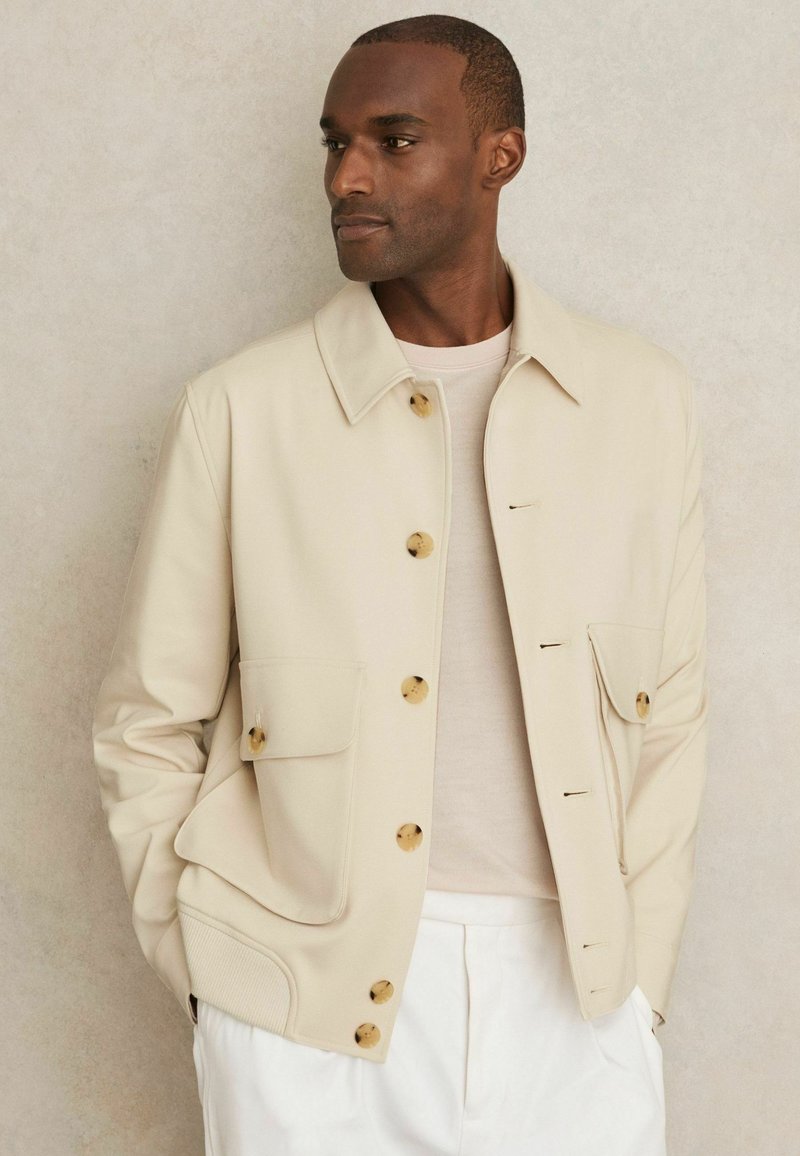 Een man met een geschoren hoofd draagt een lichtbeige knopenjack over een lichte shirt, staande tegen een neutrale achtergrond.