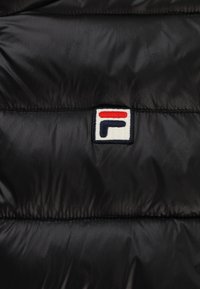 Dettaglio di un piumino nero con texture trapuntata. Sulla parte anteriore è visibile un patch del logo FILA in bianco, blu e rosso.