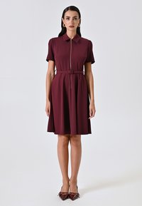 Kurzärmliges, burgunderfarbenes Kleid mit Kragen, zentralem Reißverschluss, Taillengürtel und ausgestelltem Rock. Glattes Material, knielanger Schnitt.