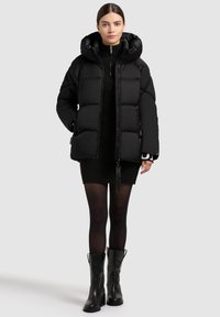 Naine kannab musta puff Jacketit koos kapuutsiga, musta soonikuga kleiti, läbipaistvaid musti sukkpükse ja musti säärtesse ulatuvaid saapaid, seistes ühetaolise taustaga.