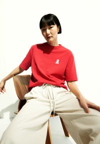 T-shirt rosso a maniche corte con logo a nastro bianco sul petto, abbinato a pantaloni beigi con coulisse. Tessuto morbido e casual; vestibilità rilassata.