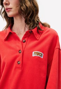 Polo-shirt en coton rouge avec un col à boutons, ornée d'un patch brodé "FRICH" en bleu et marron. Texture douce et coupe décontractée.