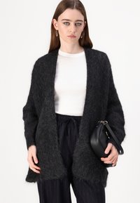 Svart, fuzzy cardigan med åpen front og lange ermer, kombinert med en hvit tettsittende topp og svarte plissert bukser. Modellen holder en svart veske.