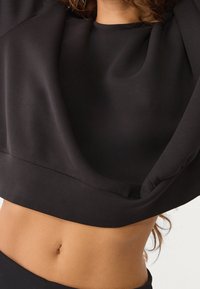 Musta lyhyt crop-top, joka on valmistettu sileästä kankaasta. Siinä on pyöreä pääntie ja pehmeä laskeutuminen pääntiellä ja hihoissa.