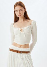 Witte lange mouw crop top met een ruches halslijn en een sleutelgat detail, gecombineerd met een witte gerimpelde rok met een bruine riem.