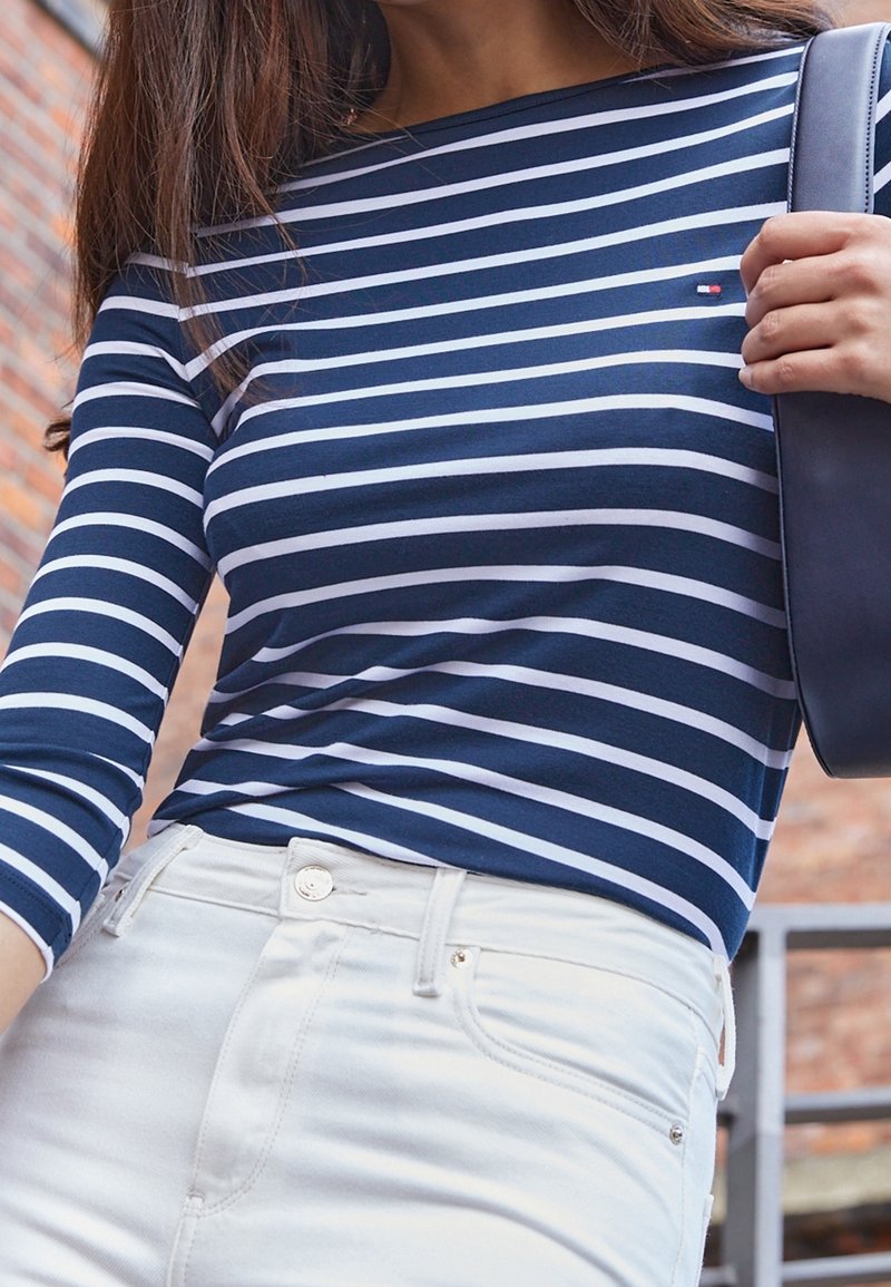 Marinblå och vit randig långärmad topp, i kombination med vita jeans med knappstängning. En svart väska hålls av modellen.