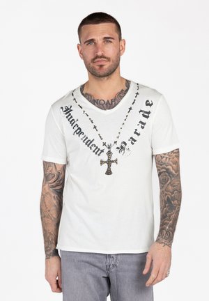 Mann mit kurzem Haar und Tattoos, trägt ein weißes T-Shirt mit einem Kreuzanhänger und dem Schriftzug "Independent Parade", steht vor weißem Hintergrund.