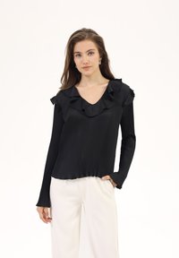 Black pleated long-sleeve top with a ruffled V-neckline, featuring a smooth texture and gathered hem. Styled with cream-colored pants.