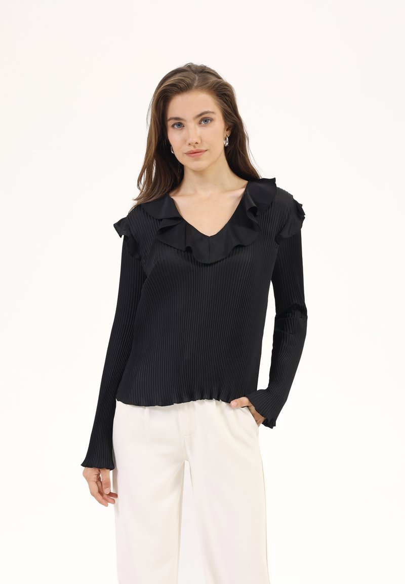 Top lungo a maniche lunghe plissonato nero con scollatura a V arricciata, caratterizzato da una texture liscia e un orlo arricciato. Abbinato a pantaloni di colore crema.