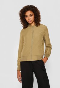 Khakifarbene Bomberjacke mit gerippten Bündchen und Saum, mit einem Frontreißverschluss und leichtem Material, kombiniert mit schwarzen Cargo-Hosen.