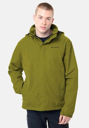 Junger Mann, der eine olivgrüne Vaude-Jacke mit Kapuze trägt, steht mit einer Hand in der Tasche vor einem hellen Hintergrund.