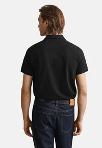 Melnā polo krekls ar apkakli, īsām piedurknēm un taisnu griezumu, kopā ar tumši zilām džinsa biksēm un melnu jostu ar ādas akcentu.