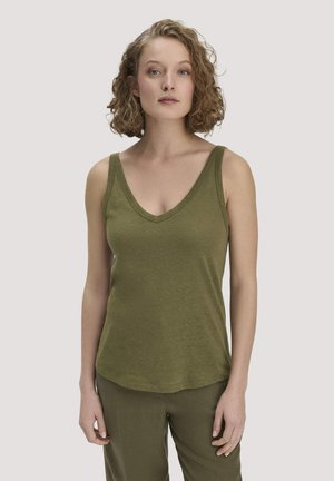 Frau mit kurzen lockigen Haaren, die ein olivgrünes ärmelloses Oberteil mit V-Ausschnitt und passende Hose trägt, stehend vor einem einfarbigen Hintergrund.