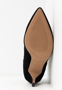 Chaussure à talons hauts en suede noir avec un bout pointu, une semelle brune texturée et un talon étroit. Nom de la marque visible sur la semelle.