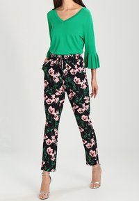 Haut vert à col en V avec manches évasées, associé à un pantalon noir à motif floral avec des fleurs roses et des feuilles vertes. Sandales argentées à talons.