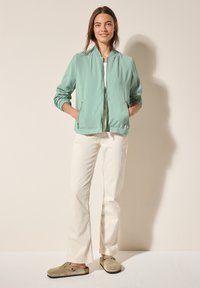 Veste bomber vert clair avec des poignets élastiques, t-shirt blanc, pantalon droit crème et chaussures à enfiler beiges avec deux trous sur les côtés.
