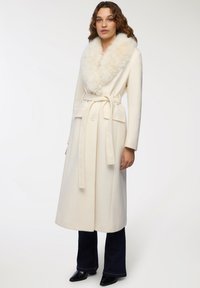 Cappotto bianco lungo con colletto in pelliccia sintetica, vita con cintura e due tasche frontali. Tessuto morbido, design lungo e vestibilità classica.