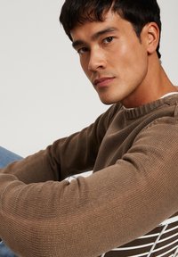 Brązowy sweter dziany z żeberkowym dekoltem i teksturowanym wzorem, noszony przez osobę, siedzącą na białym krzesle z drutu, w niebieskich dżinsach.