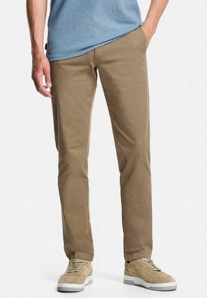 Homme avec la main dans la poche, portant un pantalon chino beige, des baskets beige en daim, et une chemise bleue à manches courtes, debout devant un fond blanc.