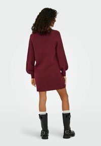 Robe pull tricotée bordeaux avec des manches longues, des poignets côtelés et une coupe décontractée, assortie à des bottes noires jusqu'aux genoux et des chaussettes blanches.
