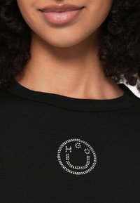 Čierne tričko s okrúhlym výstrihom, v strede zdobené grafickým motívom úsmevu z kryštálikov. Hladká textúra, priliehavý strih.