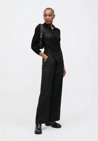 Žena nosi crnu polo majicu s dugim rukavima, široke prugaste hlače, crne cipele i torbu, stoji s jednom rukom u džepu.