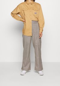 Camicia button-up beige con due tasche sul petto e logo "TOMMY", abbinata a pantaloni a gamba larga a quadri e sneakers bianche.