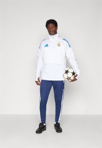 Witte sportjacket met blauwe accenten en logo, gecombineerd met blauwe broek met witte strepen. Zwarte voetbal in de hand.