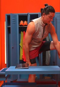 Veste grise sans manches et shorts rouges portés par un homme dans un vestiaire bleu, avec des casiers et du matériel sportif sur un fond orange vif. Tablette sur la table.