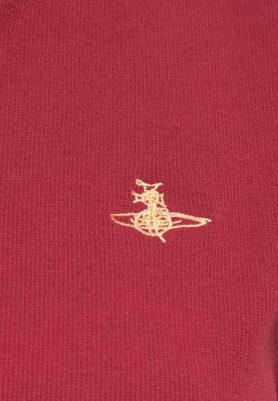 Bordo neulottu kangas, jossa on teksturoitu pinta, ja joka esittää kultaisella brodeeratulla logolla tyyliteltyä eläintä leikkisässä asennossa.