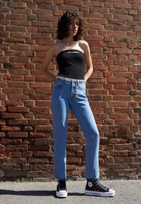 Nuori nainen, jolla on kiharat hiukset, pukeutuneena mustaan putkeen, vaaleansinisiin farkkuihin, joissa on ketjuvyö, ja mustiin korkean varren tennareihin. Hän seisoo tiiliseinää vasten.