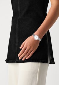 Silver analog watch with a crystal-embellished bezel, round white dial, and a decorative metal band, positioned on a hand against dark clothing.