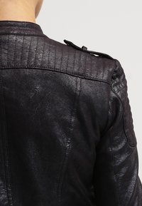 Zbliżenie na osobę w czarnej skórzanej kurtce z pikowanymi panelami na ramionach i przewiązaną paskiem z guzikami.