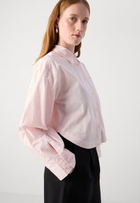 Camicia cropped rosa chiaro con colletto classico, maniche lunghe e una tasca sul petto, abbinata a pantaloni neri.