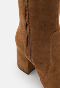 Primer plano de un tacón de bota de ante marrón con cremallera visible y detalles de costura sobre un fondo blanco.