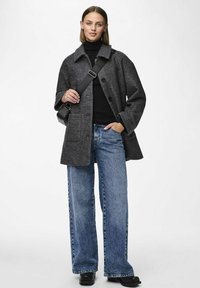 Cappotto grigio in misto lana con colletto, bottoni frontali e due tasche laterali, abbinato a jeans larghi blu a vita alta e scarpe nere.