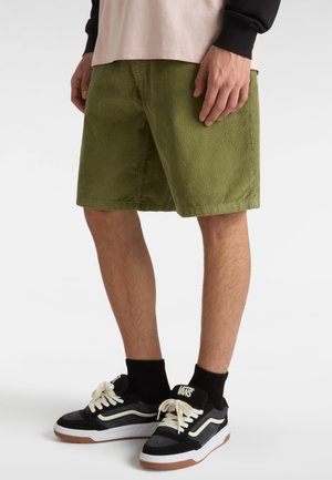 Pantaloni corti in velluto a coste verde con una texture a coste, indossati con sneaker in bianco e nero, caratterizzate da una suola bianca spessa e lacci di colore chiaro.