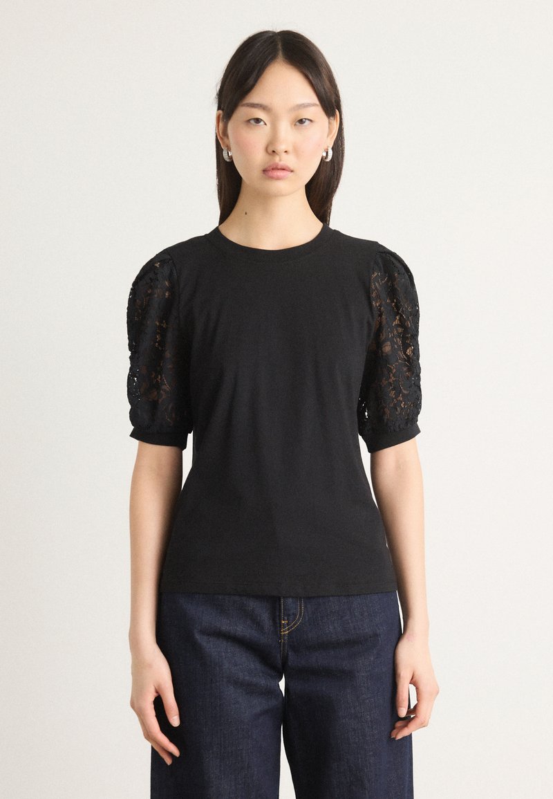 Μαύρο μπλουζάκι με κοντά, φουσκωτά μανίκια φτιαγμένο από περίπλοκο μαύρο δαντέλα. Λείος ύφασμα σώματος, εφαρμοστή σχεδίαση, σε συνδυασμό με σκούρους τζιν παντελόνες.