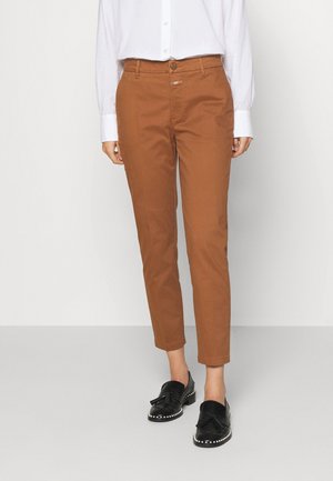 Persoon draagt roestkleurige slim-fit broek, witte blouse met lange mouwen en zwarte loafers met kwastjes en pareldetails.
