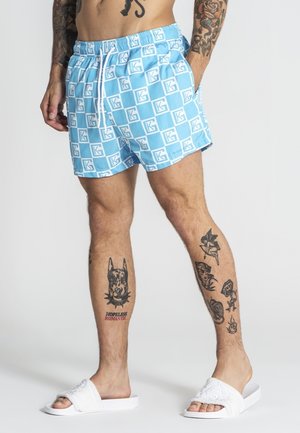 Mężczyzna w niebiesko-białych szortach kąpielowych w kratę z logo "KG", białych klapkach i tatuażami na obu nogach i ramionach, stojący na białym tle.