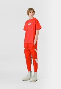 Camiseta de algodón roja y pantalones de chándal a juego con patrones de plumas blancas. El modelo lleva botas de combate crema, mostrando un ajuste relajado y elementos de diseño.