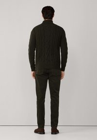 Man med mörkgrön kabelstickad tröja, matchande byxor och bruna skor som står vänd mot en enfärgad ljusgrå bakgrund.