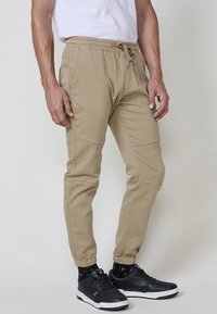 Bărbat îmbrăcat în pantaloni jogger bej cu manșete elastice, adidași negri și un tricou alb, stând pe un fundal simplu deschis.