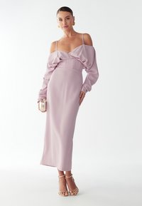 Dlouhé lila šaty s odhalenými rameny, s drapériemi a upnutým pasem. V doplnění se zlatými sandály na vysokém podpatku a malou metalickou kabelkou.