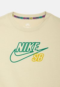 Světlobéžové tričko Nike SB s zeleným logem "NIKE" a žlutým textem "SB". Měkká tkanina s kulatým výstřihem a vzorovaným interiérovým límcem.